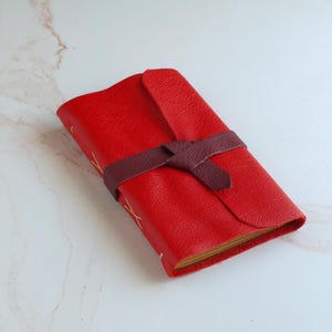 May include: A bright red leather journal with a textured surface, featuring a dark brown leather wrap-around tie closure. The spine is bound with visible light-colored stitching in an X-pattern, and the pages are visible along the side edge.