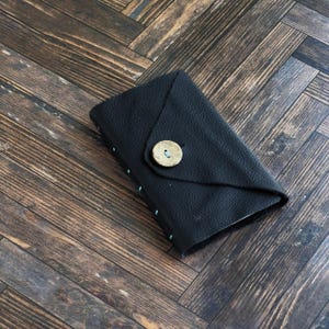 May include: A black leather pouch with a textured surface, featuring a triangular flap closure secured by a single round, light-colored wooden button. The edges of the pouch are joined with visible light blue stitching along the side.