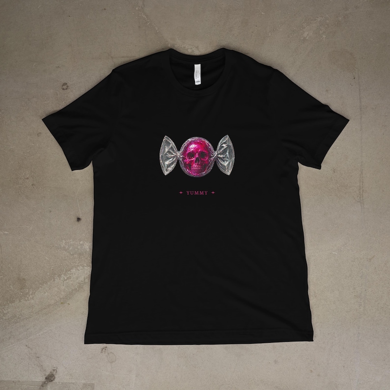 May include: Black t-shirt featuring a pink skull candy graphic. The candy is wrapped in silver foil with the word "YUMMY" printed below the graphic. The shirt is made of a soft material.