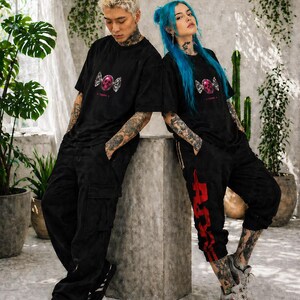 May include: Two people wearing black t-shirts with a pink skull and wing graphic. The person on the left is wearing black cargo pants and the person on the right is wearing black sweatpants with red accents. Both have tattoos and are leaning against a concrete structure.