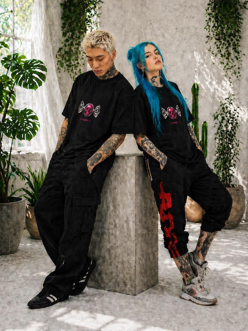 May include: Two people wearing black t-shirts with a pink skull and wing graphic. The person on the left is wearing black cargo pants and the person on the right is wearing black sweatpants with red accents. Both have tattoos and are leaning against a concrete structure.