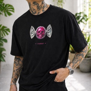 May include: Black t-shirt featuring a pink skull design wrapped in silver foil, resembling candy. The word "YUMMY" is printed below the graphic in pink. The shirt has short sleeves and a relaxed fit.