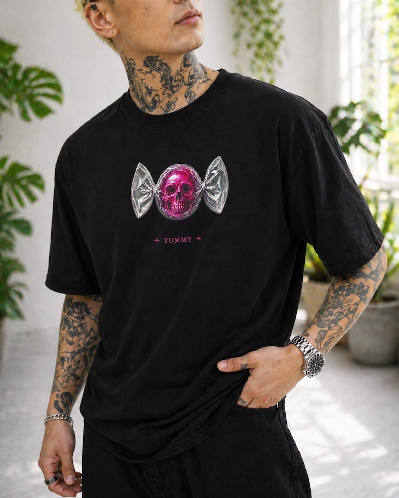 May include: Black t-shirt featuring a pink skull design wrapped in silver foil, resembling candy. The word "YUMMY" is printed below the graphic in pink. The shirt has short sleeves and a relaxed fit.