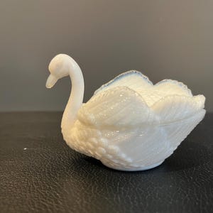 Puede incluir: Una figura de cisne blanco con un diseño detallado de plumas. El cisne tiene un cuello largo y curvado y un pico delicado. Los bordes de las alas tienen un sutil acento azul. La figura probablemente está hecha de vidrio o porcelana.