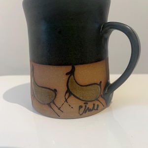 May include: A ceramic mug with a black upper section and a tan lower section. The lower section features two stylized deer in brown and green, with a signature in black. The mug has a curved handle.