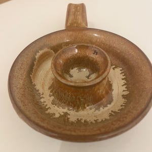 Könnte beinhalten: Ein handgefertigter Kerzenhalter aus Keramik in Braun- und Beigetönen. Er hat eine runde Basis mit einer zentralen Halterung für eine Kerze und einen Griff. Das Design wirkt rustikal und handgemacht, ideal für dekorative Zwecke.