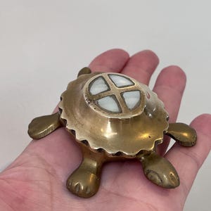 May include: A small brass turtle figurine with a scalloped shell edge. The center of the shell features a circular inlay divided into four sections by a cross-shaped brass frame, with white, smooth, opaque material filling each of the four quadrants.