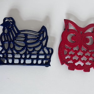 Puede incluir: Dos salvamanteles de hierro fundido: una gallina azul marino en una cesta y un búho rojo. El salvamanteles de gallina es una silueta con un diseño detallado. El salvamanteles de búho tiene un diseño recortado.