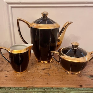 May include: A black and gold tea set comprising a teapot, creamer jug, and sugar bowl. The set has gold trim and vertical gold stripes. The teapot features a lid with a gold finial and a curved handle. The creamer and sugar bowl also have gold accents.