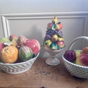 May include: Three ceramic fruit arrangements. A large white basket holds red, orange, and green ceramic fruit. A pedestal fruit sculpture features a variety of ceramic fruits. A smaller white basket holds more ceramic fruit.