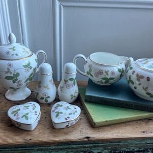 Puede incluir: Un juego de té de porcelana vintage con un diseño de fresas. El juego incluye una azucarera, una cremaera, saleros y pimenteros y joyeros en forma de corazón. Las piezas son blancas con ribetes dorados y presentan hojas verdes y fresas rosas.