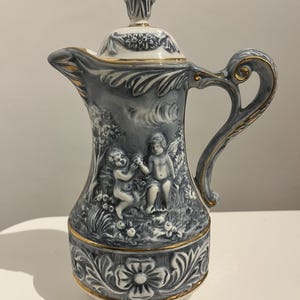 Puede incluir: Una jarra de cerámica con tapa, con una combinación de colores gris y blanco con detalles dorados. La jarra presenta un diseño en relieve detallado de querubines y motivos florales. El asa y el borde están adornados con ribetes dorados.