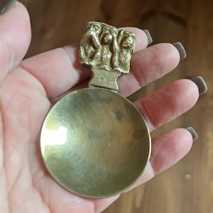 Puede incluir: Una cuchara de latón vintage con un cuenco redondo y un mango con tres figuras de monos. La cuchara tiene un acabado deslustrado, lo que sugiere antigüedad. Las figuras de monos del mango son detalladas, añadiendo un elemento decorativo al utensilio.