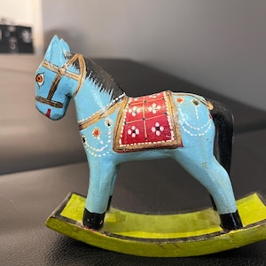 May include: A hand-painted wooden rocking horse in light blue, black, red, and yellow. The horse has a detailed saddle and bridle, with gold accents. The base is a bright yellow-green.