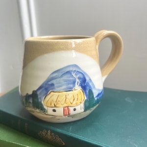Puede incluir: Una taza de cerámica pintada a mano con un paisaje. La taza presenta una parte superior beige, una sección media blanca y una montaña azul con una cabaña con techo amarillo. El asa y el borde son beige. La taza está sobre un libro verde.