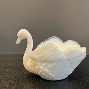 May include: A white swan-shaped trinket dish with a textured surface. The swan has a long, curved neck and detailed feather patterns. The rim of the dish has a subtle blue accent.