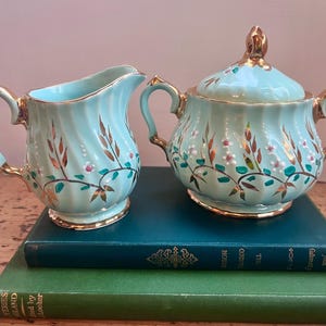 Op de afbeelding: Een vintage set van een lichtblauwe melkkan en suikerpot met deksel. De keramische stukken hebben gouden randen en bloemmotieven met bruine, groene en roze accenten. De set wordt getoond op een stapel boeken.
