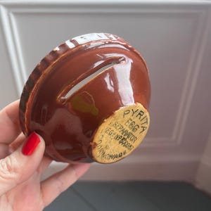 Puede incluir: Un separador de huevos de cerámica marrón con una ranura para separar las claras. El separador en forma de cuenco tiene un borde estriado y una etiqueta que dice "PYRITA EGG SEPARATOR REG. NO. 60957 COOKINGWARE."