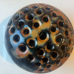 May include: A round, ceramic flower frog with numerous holes. The frog is glazed in a mix of dark blue, black, and orange hues. The holes are evenly spaced across the surface, creating a unique texture. This decorative piece is ideal for floral arrangements.
