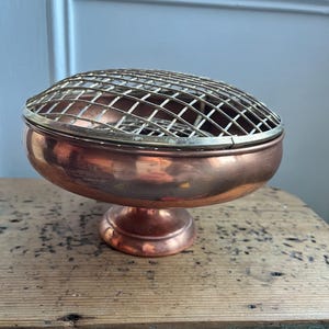 Op de afbeelding: Een koperkleurige kom met voet en een draadraster bovenop. De kom heeft een afgeronde basis en een brede, ondiepe komvorm. Het draadraster is gemaakt van een lichter gekleurd metaal, wat een contrast creëert met het koper. De kom staat op een houten oppervlak.