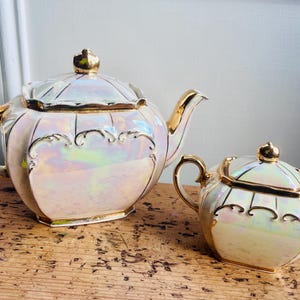 May include: A pearlescent tea set with gold trim. The set includes a teapot with a lid and a sugar bowl with a lid. The teapot has a curved spout and handle. The sugar bowl has two handles. Both pieces have decorative detailing.