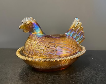 May include: An iridescent glass hen-shaped dish with a lid, sitting on a round base. The hen is a mix of gold, orange, and blue hues. The base has a beaded edge, and the hen's feathers are detailed.