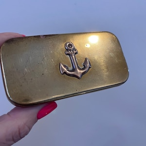 May include: A rectangular brass-coloured metal case with rounded corners, featuring a centred, raised silver-toned anchor emblem on the lid. The surface of the case shows signs of wear, including light scratches and a dull, aged patina finish.
