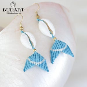 Könnte beinhalten: Handgefertigte Makramee-Ohrringe mit weißen Muscheln und hellblauen und weißen Makramee-Meerjungfrauenschwanz-Designs. Die Ohrringe haben goldfarbene Haken und Akzente. Das Budart-Logo ist sichtbar.