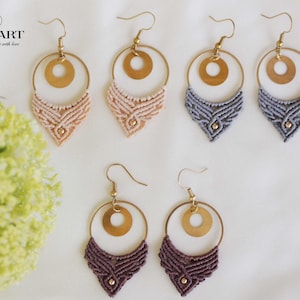 Könnte beinhalten: Eine Sammlung handgefertigter Makramee-Ohrringe in verschiedenen Farben. Jeder Ohrring hat einen goldfarbenen Reifen mit kreisförmigem Akzent und ein gewebtes Makramee-Design in Beige, Grau und Lila. Die Ohrringe werden auf einer weißen Oberfläche präsentiert.