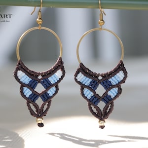 May include: A pair of handmade macrame earrings. Each earring features a gold-colored hoop with a woven design in shades of brown, white, and blue. The earrings have a small gold bead at the bottom and are designed for pierced ears.