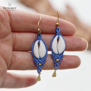 Può includere: Un paio di orecchini macramè blu fatti a mano con conchiglie bianche, dettagli dorati e ciondoli triangolari. Gli orecchini hanno ganci per le orecchie dorati. Il marchio "BUDART" è visibile.