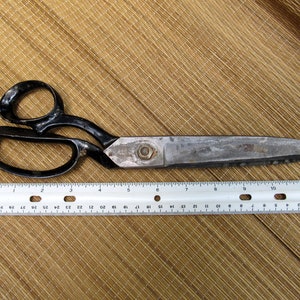 May include: Large, vintage metal shears with black handles and a silver blade, next to a ruler. The shears are approximately 11 inches long. The ruler shows measurements in inches and millimeters. The shears are on a textured, tan surface.