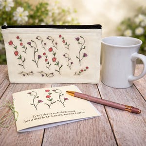 May include: A cream-colored zippered pouch, white mug, pen, and a small notebook are arranged on a wooden surface. The pouch and notebook feature floral designs. The notebook reads, "Every day is a new beginning. Take a deep breath, smile, and start again."