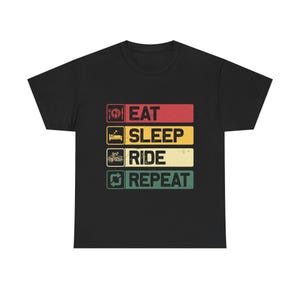 Op de afbeelding: Zwart T-shirt met een retro design. De tekst "EAT SLEEP RIDE REPEAT" is gestapeld op verschillende gekleurde rechthoeken. Iconen zijn een vork en mes, een bed, een motorfiets en een herhaalsymbool, wat een vintage uitstraling creëert.