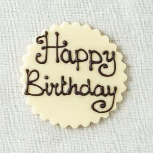 May include: A circular biscuit with a decorative, scalloped edge. The words "Happy Birthday" are written in dark brown icing in a cursive font. The biscuit is set against a textured, off-white background.