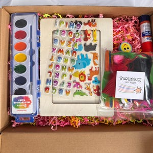 May include: A craft box filled with art supplies. Includes a watercolor paint set, wooden frame with alphabet and animal stickers, glue stick, and a bag of colorful feathers. The box is lined with pink shredded paper.