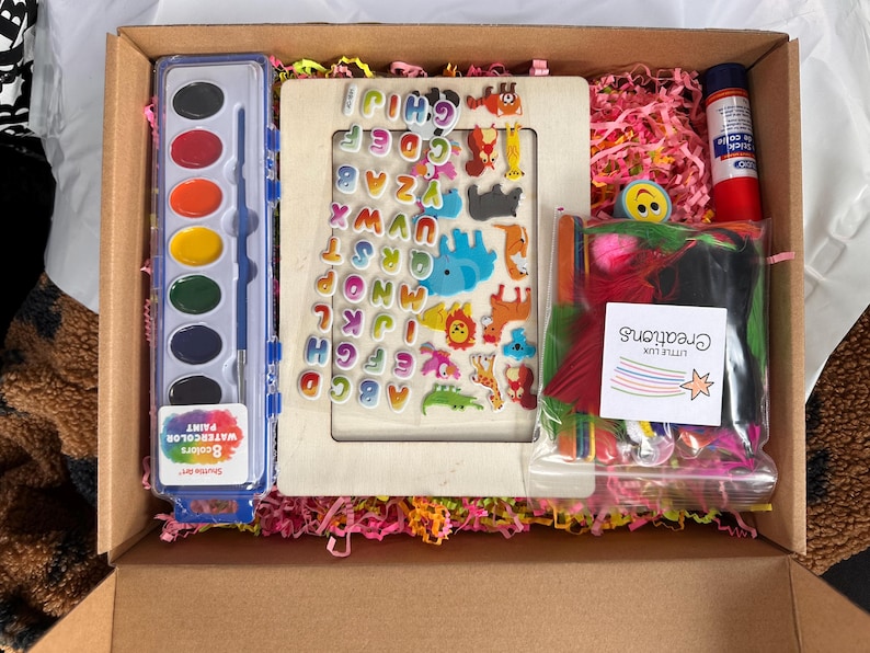 May include: A craft box filled with art supplies. Includes a watercolor paint set, wooden frame with alphabet and animal stickers, glue stick, and a bag of colorful feathers. The box is lined with pink shredded paper.