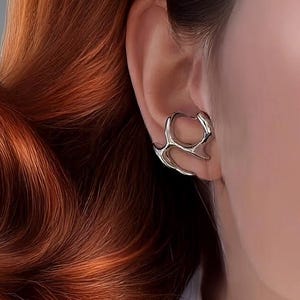 Może przedstawiać: Srebrny nausznik o otwartym, organicznym wzorze jest noszony na uchu. Nausznik jest osadzony na tle żywych, kasztanowych włosów. Włosy są ułożone w miękkie fale i widoczna jest biała koszula.