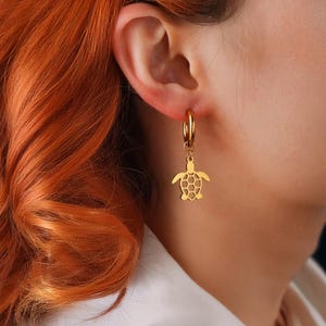 May include: Gold-coloured hoop earrings featuring a turtle charm. The charm has a detailed shell design. The earrings are displayed on a person's ear, with the focus on the jewellery.