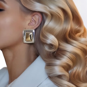 May include: Silver-toned square earrings with a gold-colored center. The earrings are worn on a person with wavy blonde hair and a white collared shirt. The earrings have a geometric design.