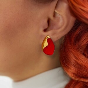 Puede incluir: Un par de pendientes de declaración dorados y rojos. Los pendientes tienen forma de lágrima con un contorno dorado y un centro rojo.