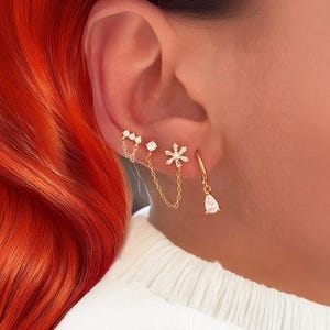 Op de afbeelding: Een verzameling gouden oorbellen met heldere edelstenen. De oorbellen omvatten een kleine oorring met een druppelvormige edelsteen, een ketting met een ronde edelsteen en een ster, en een dubbele ketting met hartvormige edelstenen.