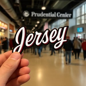May include: A black, white, and red sticker with the word "Jersey" in a cursive font. The sticker is held in front of a blurred background of a crowded indoor space with a sign that reads "Prudential Center".