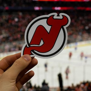 May include: A red and white sticker featuring the New Jersey Devils logo, a stylized "N" with devil horns, held in front of a hockey game. The logo is set against a white background with a black outline.