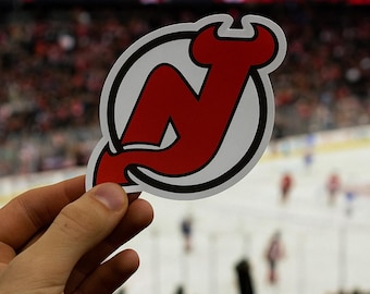 May include: A red and white sticker featuring the New Jersey Devils logo, a stylized "N" with devil horns, held in front of a hockey game. The logo is set against a white background with a black outline.