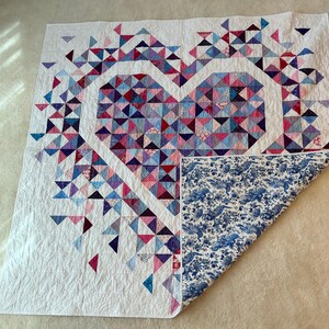 Puede incluir: Un edred&oacute;n hecho a mano con un dise&ntilde;o de coraz&oacute;n hecho de tri&aacute;ngulos coloridos en tonos de rosa, morado y azul. El edred&oacute;n tiene un fondo blanco y un reverso con estampado floral azul.