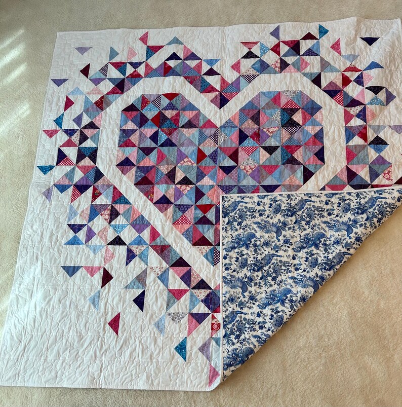 Puede incluir: Un edred&oacute;n hecho a mano con un dise&ntilde;o de coraz&oacute;n hecho de tri&aacute;ngulos coloridos en tonos de rosa, morado y azul. El edred&oacute;n tiene un fondo blanco y un reverso con estampado floral azul.