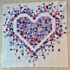 Puede incluir: Un edred&oacute;n blanco con un dise&ntilde;o de coraz&oacute;n hecho de piezas de tela triangulares de colores. El coraz&oacute;n est&aacute; delineado en blanco, con tri&aacute;ngulos rosas, azules y morados que llenan el interior. El edred&oacute;n tiene una apariencia suave y texturizada.