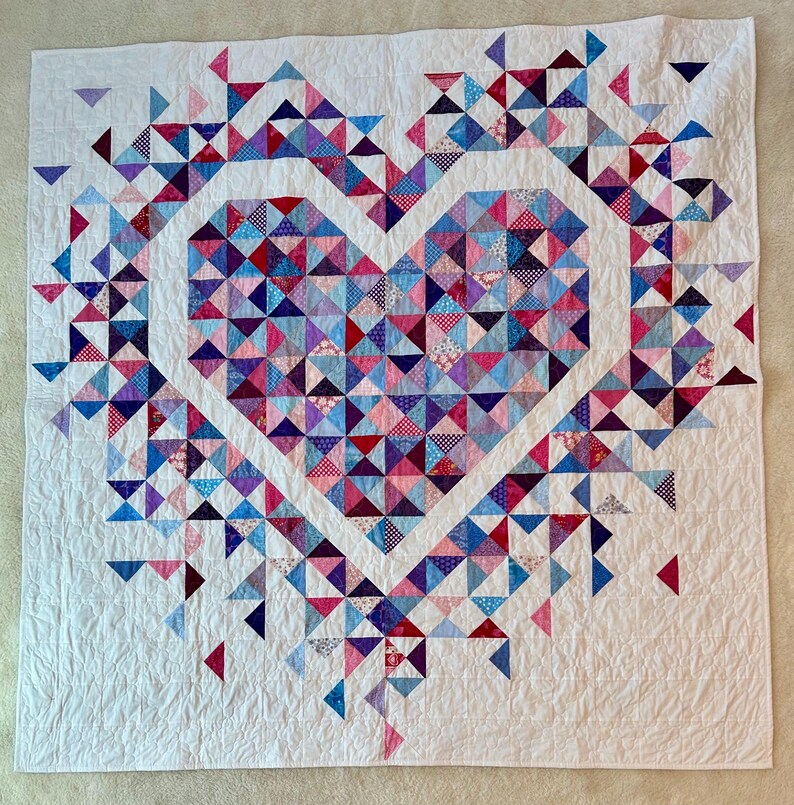 Puede incluir: Un edred&oacute;n blanco con un dise&ntilde;o de coraz&oacute;n hecho de piezas de tela triangulares de colores. El coraz&oacute;n est&aacute; delineado en blanco, con tri&aacute;ngulos rosas, azules y morados que llenan el interior. El edred&oacute;n tiene una apariencia suave y texturizada.