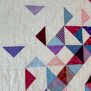 Puede incluir: Primer plano de un edred&oacute;n hecho a mano con un fondo blanco y patrones triangulares coloridos. Los tri&aacute;ngulos son en tonos de azul, morado, rosa y rojo, con varios patrones. El edred&oacute;n est&aacute; cosido a m&aacute;quina con un patr&oacute;n ondulado.